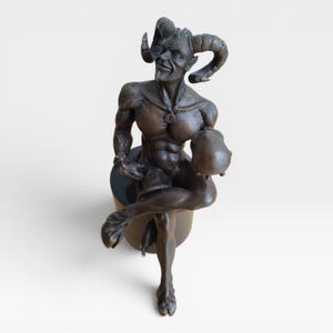 May include: A bronze-toned sculpture of a satyr with large curved horns, pointed ears, and a goatee. The figure has a muscular torso, cloven hooves, and a tail, sitting on a cylindrical base while holding a round object in one hand.