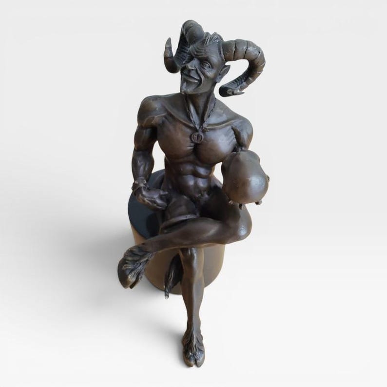 May include: A bronze-toned sculpture of a satyr with large curved horns, pointed ears, and a goatee. The figure has a muscular torso, cloven hooves, and a tail, sitting on a cylindrical base while holding a round object in one hand.