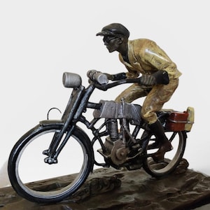 以下が含まれることがあります： ヴィンテージバイクに乗る人物のブロンズ像。ライダーは帽子と黄色のジャケットを着用しています。バイクは黒とシルバーのアクセント、赤い収納ボックス付き。彫刻は暗い台座に取り付けられています。