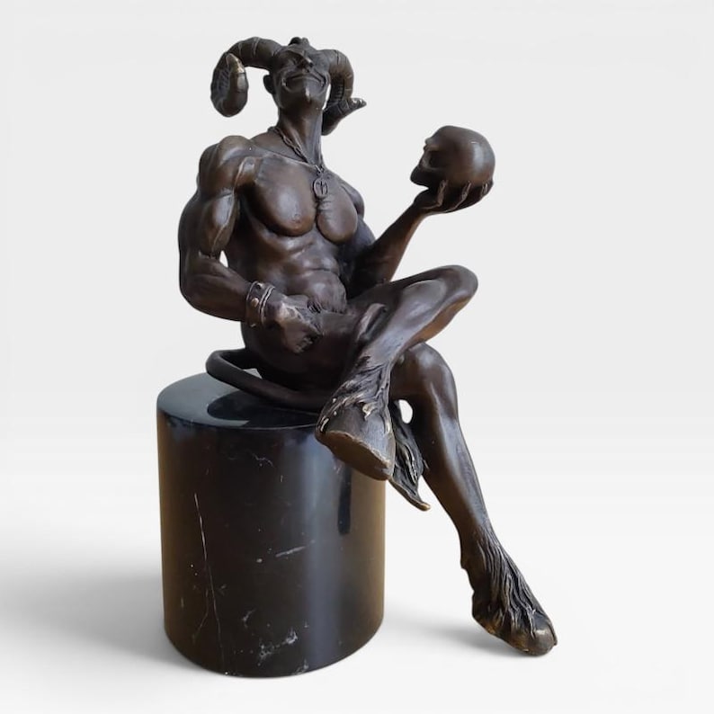 May include: A bronze-toned sculpture of a horned, muscular figure with cloven hooves sitting on a cylindrical black marble pedestal. The figure wears a necklace and wristband, and holds a human skull in one hand while looking forward.