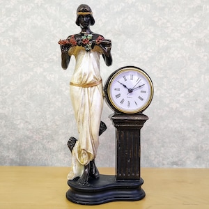 Puede incluir: Una figura de mujer en blanco y negro sosteniendo una corona de flores se encuentra sobre una base con una esfera de reloj. La esfera del reloj tiene números romanos y agujas doradas.