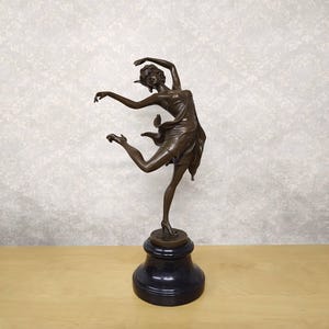 May include: Bronze sculpture of a dancing figure in a flowing dress, poised mid-leap. The sculpture is mounted on a black, cylindrical base. The dancer's arms are outstretched, and her hair is styled up.