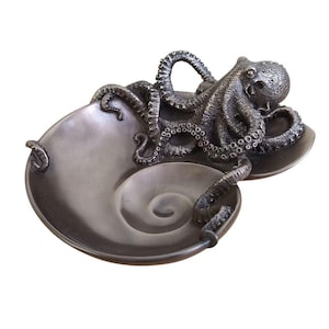 Può includere: Una scultura di un polpo color argento con tentacoli dettagliati, che tiene un piatto a forma di conchiglia. Il polpo è avvolto attorno al piatto, con i suoi tentacoli che si estendono verso l'esterno.