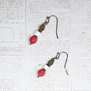 Può includere: Orecchini pendenti fatti a mano con perline rosse, bianche e verde oliva. Gli orecchini hanno un'estetica vintage, con un gancio color argento. Lo sfondo è una pagina di giornale vintage. Un accessorio unico.