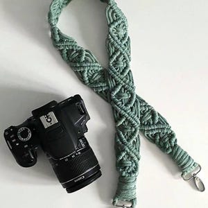 Puede incluir: Una correa de cámara de macramé verde salvia con un diseño tejido. La correa se combina con una cámara Canon negra. La correa tiene clips metálicos plateados para la fijación. La lente de la cámara tiene la palabra "Canon" impresa.