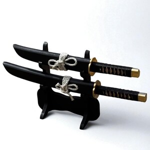 May include: Two black decorative tanto knives with gold accents and wrapped handles are displayed on a black stand. Each knife is secured with a white rope. The blades are curved and the stand is minimalist.