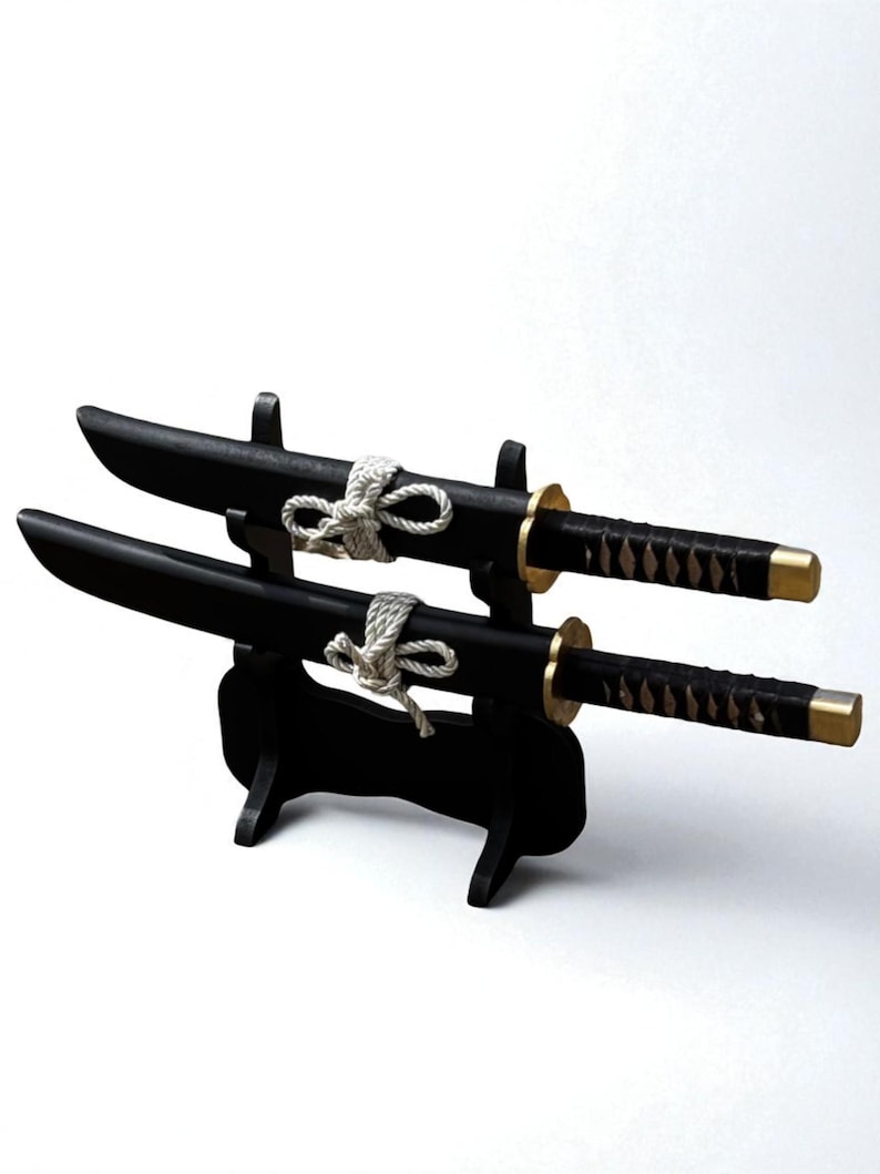 May include: Two black decorative tanto knives with gold accents and wrapped handles are displayed on a black stand. Each knife is secured with a white rope. The blades are curved and the stand is minimalist.
