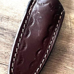 May include: A handcrafted knife with a blue and white handle and a dark brown leather sheath. The sheath is stitched with white thread and features an embossed design. The knife is designed for outdoor use.