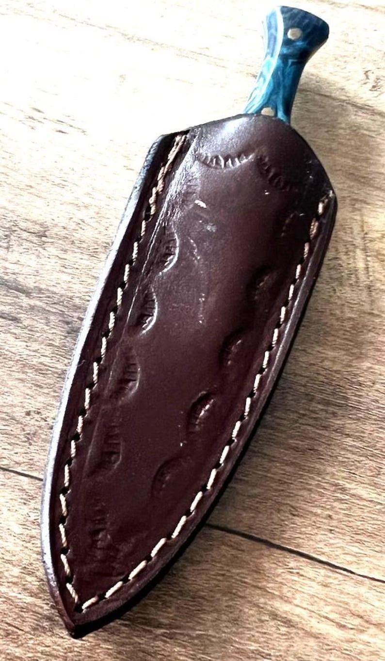 May include: A handcrafted knife with a blue and white handle and a dark brown leather sheath. The sheath is stitched with white thread and features an embossed design. The knife is designed for outdoor use.