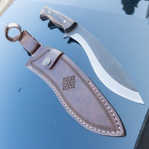 May include: A brown leather knife sheath with a decorative stitch pattern and a floral design. The sheath is for a large, curved knife with a wooden handle.
