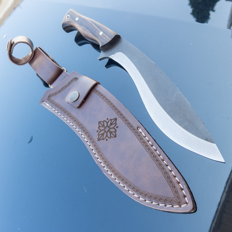 May include: A brown leather knife sheath with a decorative stitch pattern and a floral design. The sheath is for a large, curved knife with a wooden handle.