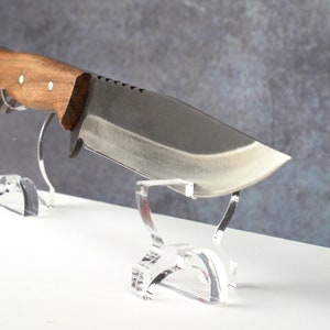May include: A large, fixed blade knife with a brown wooden handle is displayed on a clear acrylic stand. The knife has a serrated edge and a slightly curved blade.