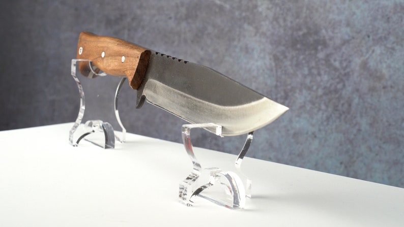 May include: A large, fixed blade knife with a brown wooden handle is displayed on a clear acrylic stand. The knife has a serrated edge and a slightly curved blade.