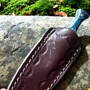 May include: A handmade knife with a blue handle and a brown leather sheath. The sheath is stitched with white thread and has a decorative pattern. The knife is resting on a stone surface with green moss in the background.