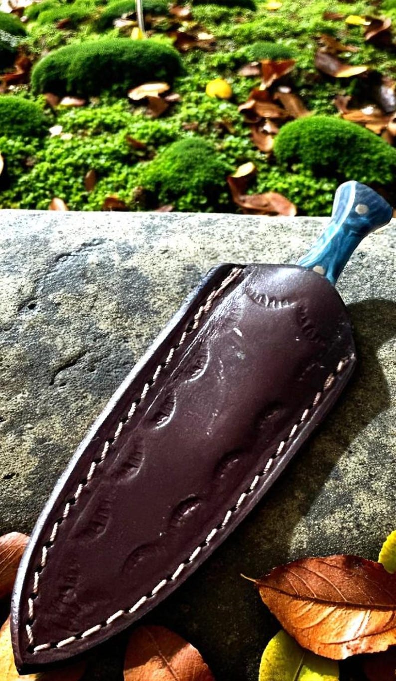 May include: A handmade knife with a blue handle and a brown leather sheath. The sheath is stitched with white thread and has a decorative pattern. The knife is resting on a stone surface with green moss in the background.