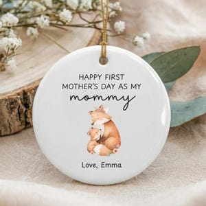May include: White ceramic ornament with gold string for hanging. The ornament features the text "HAPPY FIRST MOTHER'S DAY AS MY mommy" and a watercolor illustration of two foxes. The bottom of the ornament reads "Love, Emma".