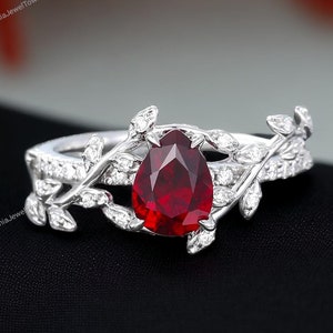Peut inclure: Une bague en argent avec une pierre précieuse en rubis de forme poire et un motif délicat de feuilles. La bague est ornée d'une rangée de petits diamants de chaque côté du rubis.