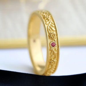 May include: A gold ring with intricate floral and leaf patterns. A small, round, red gemstone is set into the design. The ring is displayed on a black and white background, highlighting the ring's details.