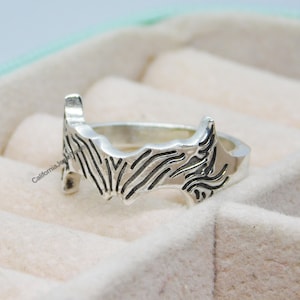 以下が含まれることがあります： 州の輪郭の細かなデザインが施された銀の指輪。白いベルベットの指輪箱に展示されています。