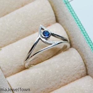 以下が含まれることがあります： 三角形のデザインに青い宝石がセットされた銀の指輪。指輪は白い指輪の箱に入っています。