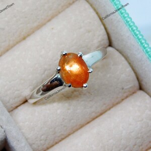 Puede incluir: Un anillo de plata con una piedra preciosa de piedra de sol naranja. El anillo está en una caja de anillos.