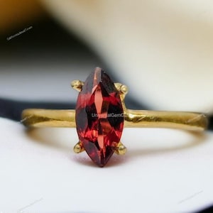 May include: A gold ring with a marquise-shaped red gemstone. The ring is set on a white surface.