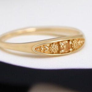 Könnte beinhalten: Ein goldener Ring mit einem ovalen Design mit floralen und geometrischen Mustern. Der Ring ist vor einem weißen Hintergrund mit einem schwarzen Band platziert, wodurch ein Kontrast entsteht, der die Details des Rings hervorhebt. Das Design des Rings ist auf dem Band zentriert.
