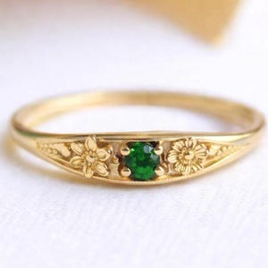 May include: A gold ring with a small, round, emerald-green gemstone at the center. The ring features floral designs on either side of the gemstone. The ring is set against a white background, with a soft, cream-colored fabric in the upper left corner.