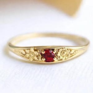 May include: A gold ring with a small, square, red gemstone at the center. The ring features floral designs on either side of the gemstone. The ring is set against a white background.