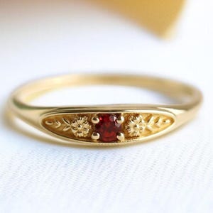 May include: A gold ring with a central red gemstone. The ring has floral and leaf patterns on the band. The ring is set against a white background with a soft, blurred backdrop.