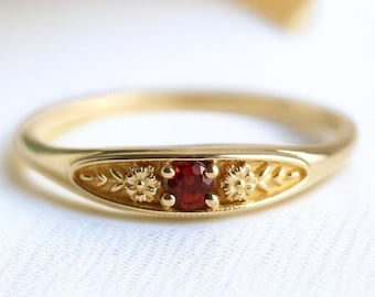 May include: A gold ring with a central red gemstone. The ring has floral and leaf patterns on the band. The ring is set against a white background with a soft, blurred backdrop.