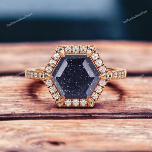 May include: A hexagonal engagement ring with a blue starburst gemstone surrounded by a halo of small diamonds. The ring is set in rose gold.