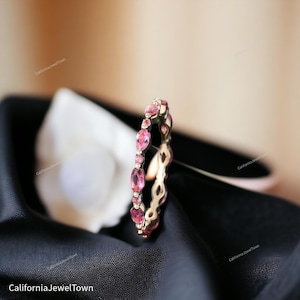 Puede incluir: Un delicado anillo de banda de oro con una fila de piedras ovaladas de color rosa. El anillo está sobre un fondo de tela negra.