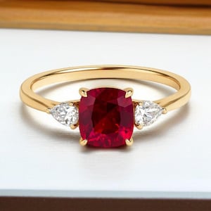 May include: A gold ring featuring a central cushion-cut deep red gemstone flanked by two pear-cut clear white gemstones. The band is polished yellow gold, creating a three-stone design that highlights the contrast between the red and white stones.