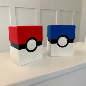 May include: Two Pokeball-themed storage boxes, one red and one blue, with a white base and a black ring around the center. The center of each box has a white circle.