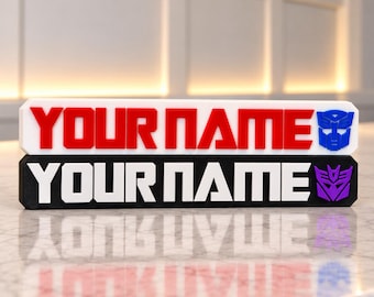 May include: Two rectangular 3D-printed nameplates. The top white plate features red text reading YOUR NAME and a blue Autobot logo. The bottom black plate features white text reading YOUR NAME and a purple Decepticon logo.