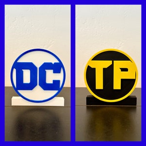 May include: Two white round plaques with black bases. The plaque on the left has a blue "DC" logo. The plaque on the right has a yellow "TP" logo.