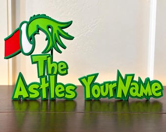 May include: A green, red, and white 3D sign featuring a stylized hand wearing a Santa hat above the text The Astles YourName. The letters are in a bold, jagged font with a layered appearance, standing upright on a flat surface.