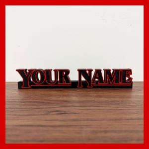 Può includere: Un cartello nero e rosso con la scritta "YOUR NAME". Le lettere sono nere con un contorno rosso. Il cartello è su una superficie di legno marrone scuro, con uno sfondo bianco.