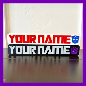 Può includere: Un'etichetta per nomi bianca e nera con il testo "YOUR NAME" in lettere rosse e argentate. L'etichetta per nomi ha un logo Transformers su ogni lato.