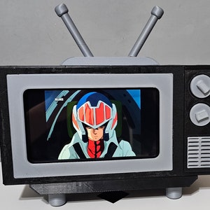 May include: A miniature black and gray television with a screen displaying an anime scene. The TV has a retro design with two gray antennas, a gray frame around the screen, and two gray knobs. The screen shows a person in a red and blue helmet.