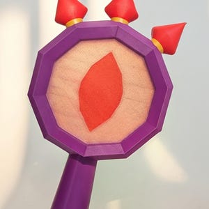 Könnte beinhalten: Eine violette achteckige Requisite mit Griff, die in der Mitte einen hellpfirsichfarbenen Stoff mit einem roten rautenförmigen Aufnäher aufweist. Drei rote kegelförmige Spitzen mit gelben Sockeln sind am oberen Rand des achteckigen Rahmens befestigt.