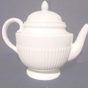 May include: A white ceramic teapot with a lid and a curved handle. The teapot has a ribbed design on the body and a decorative rope-like detail around the lid. The spout is curved and the teapot sits on a small, round base.