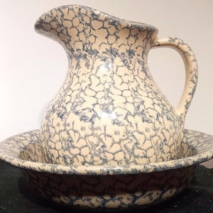 May include: A vintage ceramic pitcher and bowl set. The pitcher has a curved handle and spout. Both pieces are cream-colored with a blue, web-like pattern. The bowl sits beneath the pitcher.