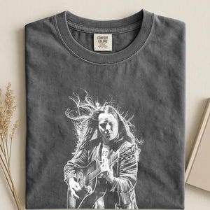 Può includere: Una t-shirt a girocollo grigio antracite con una stampa grafica bianca di una persona con i capelli lunghi che suona una chitarra acustica. L'etichetta Comfort Colors sul collo indica la taglia small, prodotta in Honduras.