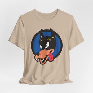 May include: A tan t-shirt featuring a cartoon wolf graphic. The wolf is black with a red tongue and blue eyes. The wolf is inside a blue circle with a black outline.