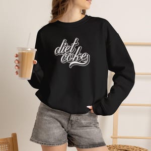 Może przedstawiać: Czarna bluza z długim rękawem z białym napisem Diet Coke stylizowaną czcionką na piersi. Ubiór jest zestawiony z szarymi szortami dżinsowymi, tworząc swobodny wygląd podczas trzymania przezroczystego plastikowego kubka ze słomką.