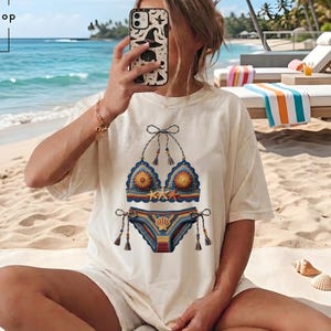 May include: An off-white short-sleeve t-shirt featuring a graphic print of a blue, orange, and yellow crochet-style bikini set with starfish and seashell accents. The text BrgBangShop is visible in the upper left corner of the image.
