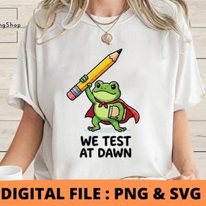 Può includere: Una t-shirt bianca con la grafica di una rana verde che indossa un mantello rosso, tiene una grande matita gialla sopra la testa e un libro sotto il braccio. Sotto la rana, il testo recita WE TEST AT DAWN in grassetto nero maiuscolo.
