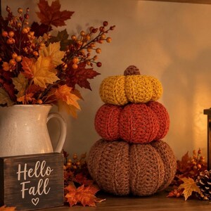 May include: A stack of three crocheted pumpkins in yellow, orange, and brown, topped with a small brown stem. Next to the pumpkins is a dark wooden sign with the white text "Hello Fall" and a heart symbol, alongside autumn leaves.